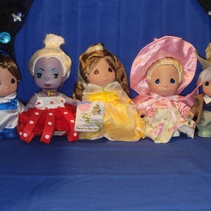 May include: Five Precious Moments dolls are displayed on a blue surface. The dolls are dressed in various outfits, including a blue and white outfit, a red and white polka-dot dress, a yellow gown, a pink floral dress, and a green outfit with a hat.