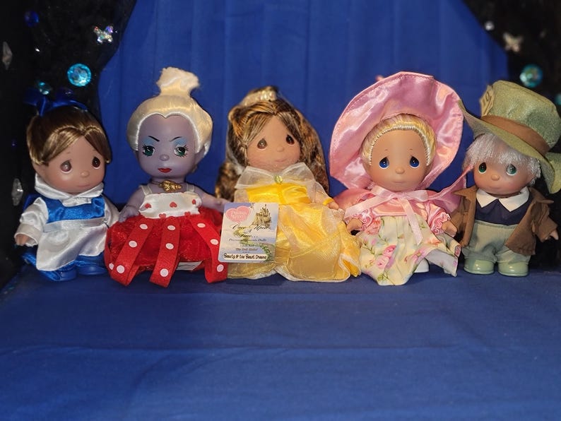 May include: Five Precious Moments dolls are displayed on a blue surface. The dolls are dressed in various outfits, including a blue and white outfit, a red and white polka-dot dress, a yellow gown, a pink floral dress, and a green outfit with a hat.