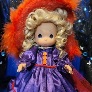 May include: A Precious Moments figurine of a girl with blonde curly hair, wearing a purple dress with orange buttons and trim. She is wearing a large orange hat with feather trim. The background is dark blue with sparkling accents.