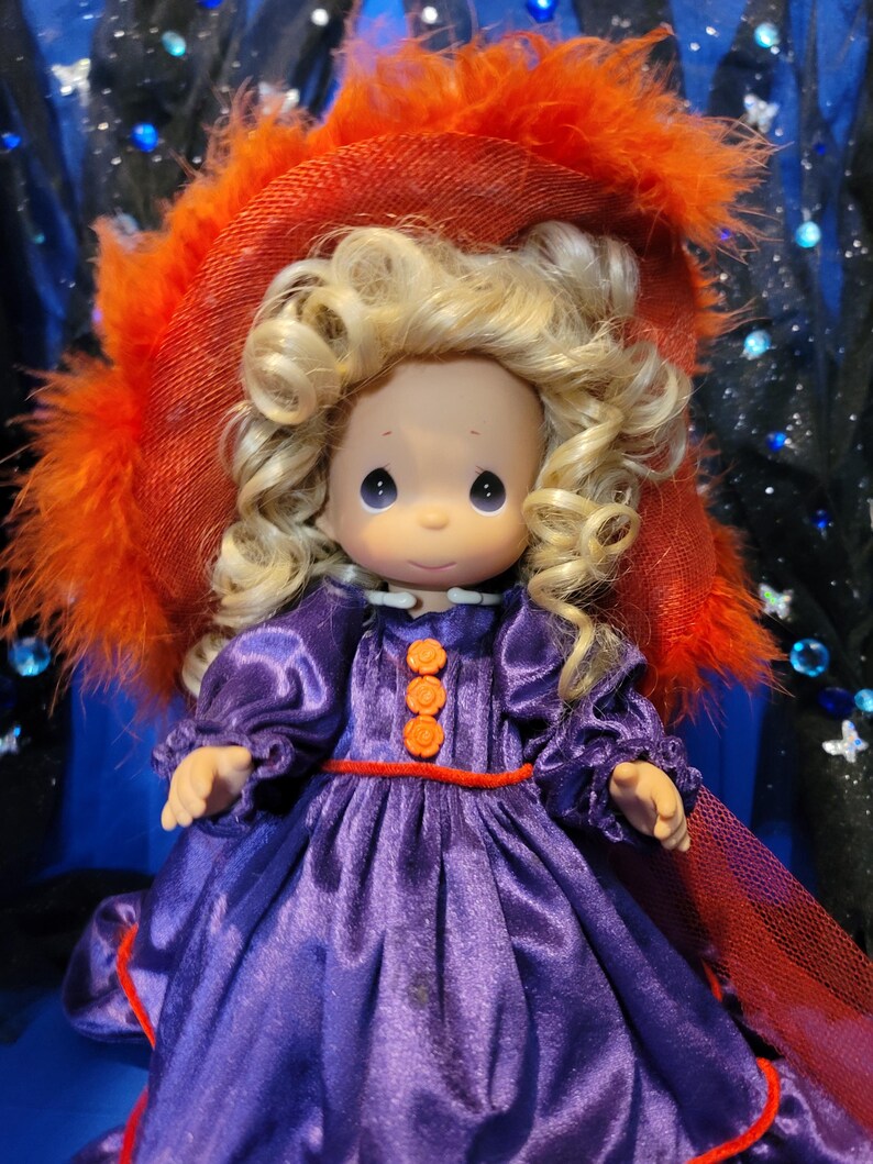 May include: A Precious Moments figurine of a girl with blonde curly hair, wearing a purple dress with orange buttons and trim. She is wearing a large orange hat with feather trim. The background is dark blue with sparkling accents.