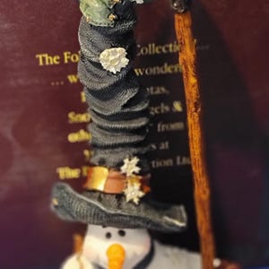 K&ouml;nnte beinhalten: Eine skurrile Schneemannfigur mit schwarzem Hut, oranger Karottennase und gestreifter Krawatte. Der Schneemann h&auml;lt einen Besen und ist mit dekorativen Akzenten verziert. Teil einer Sammlung.