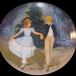 May include: A decorative plate featuring a painted scene of a young couple dancing near a Christmas tree. The girl wears a light blue dress, and the boy is in a black jacket and tan pants. The plate has a black border and the text "MoneySavingDeals58".