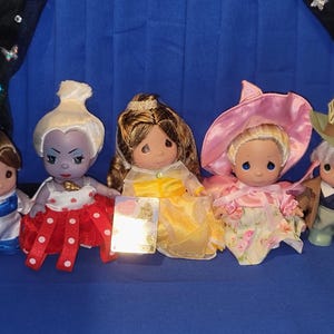 May include: A collection of Precious Moments figurines featuring various characters. The figurines are dressed in colorful outfits, including a blue and white dress, a red dress with white polka dots, and a yellow dress. Some figurines wear hats.