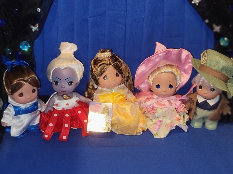 May include: A collection of Precious Moments figurines featuring various characters. The figurines are dressed in colorful outfits, including a blue and white dress, a red dress with white polka dots, and a yellow dress. Some figurines wear hats.