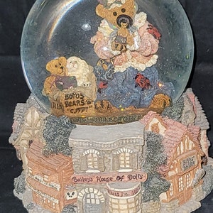 May include: A snow globe featuring a teddy bear family inside a glass sphere. The base is a detailed village scene with buildings and the text "Bailey's House of Dolls" and "Boyds Bears". The bears are holding a baby bear.