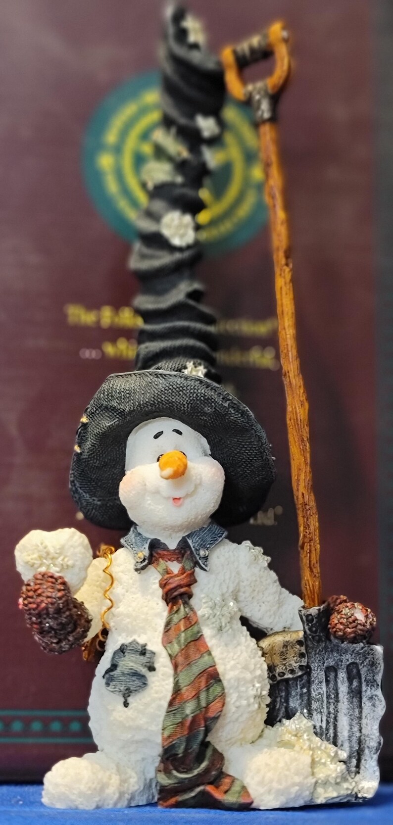 K&ouml;nnte beinhalten: Eine skurrile Schneemannfigur mit schwarzem Hut, bunter gestreifter Krawatte und Jeansjacke. Der Schneemann h&auml;lt eine Schaufel und einen Schneeball. Eine festliche Winterdekoration.