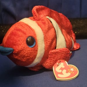 May include: A plush clownfish toy in vibrant red with white stripes and blue accents. The toy has large blue eyes and a blue mouth. A heart-shaped tag is attached to the toy.