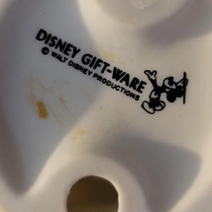 May include: Close-up of a white ceramic figurine base with the text "DISNEY GIFT-WARE" and "WALT DISNEY PRODUCTIONS" in black. A black and white Mickey Mouse graphic is also present. The figurine has a small, circular hole.