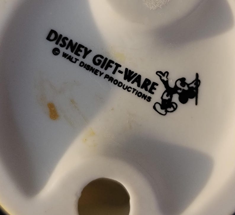 May include: Close-up of a white ceramic figurine base with the text "DISNEY GIFT-WARE" and "WALT DISNEY PRODUCTIONS" in black. A black and white Mickey Mouse graphic is also present. The figurine has a small, circular hole.