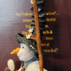 K&ouml;nnte beinhalten: Eine skurrile Schneemannfigur mit Karottennase, die einen schwarzen Hut und einen Schal tr&auml;gt. Der Schneemann h&auml;lt einen Holzstab und einen kleinen Gegenstand. Die Figur hat ein strukturiertes, schneegleiches Aussehen und ist Teil einer Sammlung.