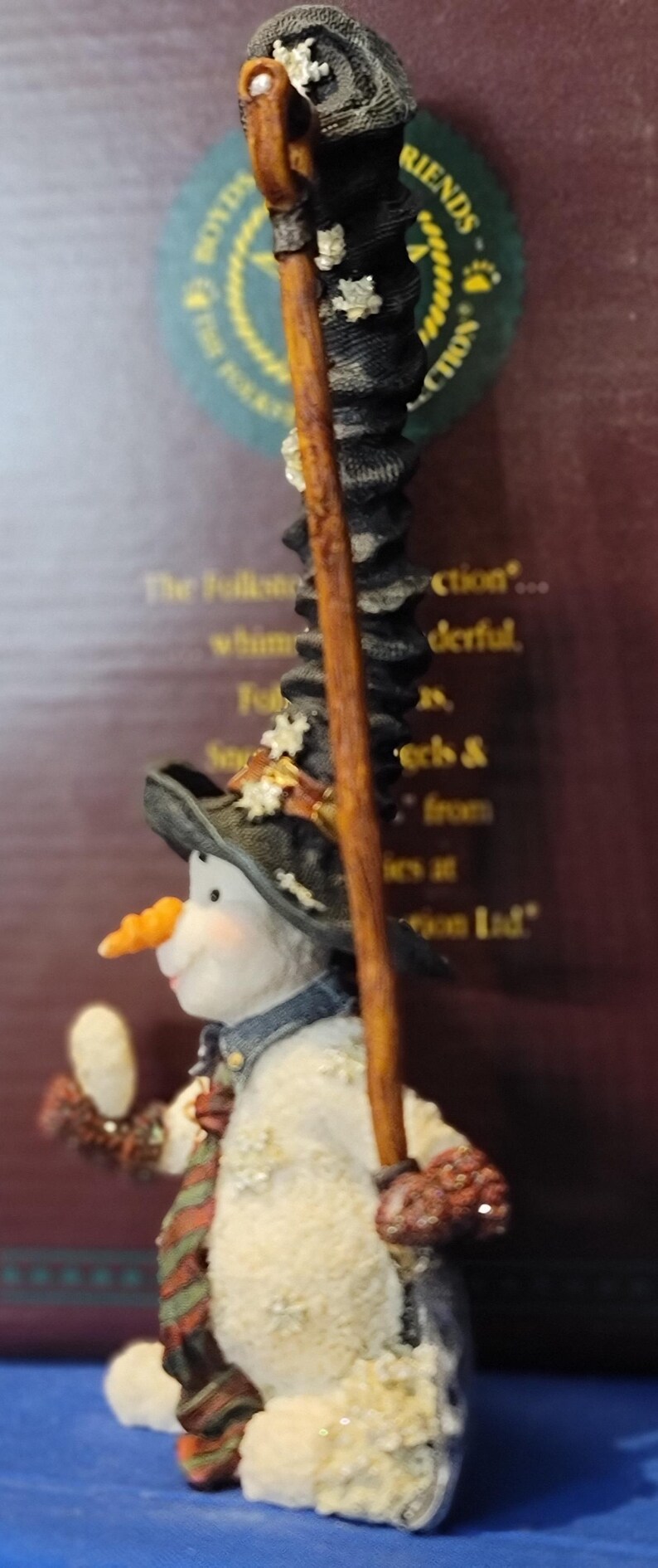 K&ouml;nnte beinhalten: Eine skurrile Schneemannfigur mit Karottennase, die einen schwarzen Hut und einen Schal tr&auml;gt. Der Schneemann h&auml;lt einen Holzstab und einen kleinen Gegenstand. Die Figur hat ein strukturiertes, schneegleiches Aussehen und ist Teil einer Sammlung.