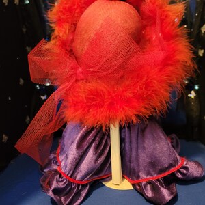 May include: A red feathered hat with a matching veil sits atop a mannequin head. The hat is paired with a purple velvet dress with red trim. The ensemble is displayed on a white stand against a blue backdrop.