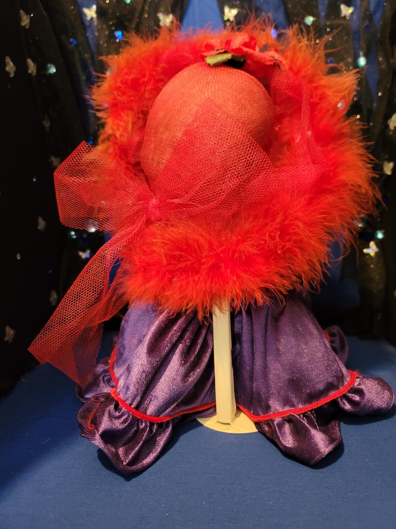 May include: A red feathered hat with a matching veil sits atop a mannequin head. The hat is paired with a purple velvet dress with red trim. The ensemble is displayed on a white stand against a blue backdrop.