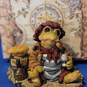 May include: A whimsical figurine of a frog dressed as a wine connoisseur, wearing a brown hat and jacket, holding a wine glass and a cigar. The frog is seated on a stone base with a barrel and bottle labeled "Old Toad". The figurine is part of "The Wee Folkstones" collection.