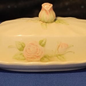 May include: A cream-colored ceramic butter dish with a rose-shaped knob, a matching oval serving plate, and two salt and pepper shakers. Each piece is decorated with pink roses and green leaves.