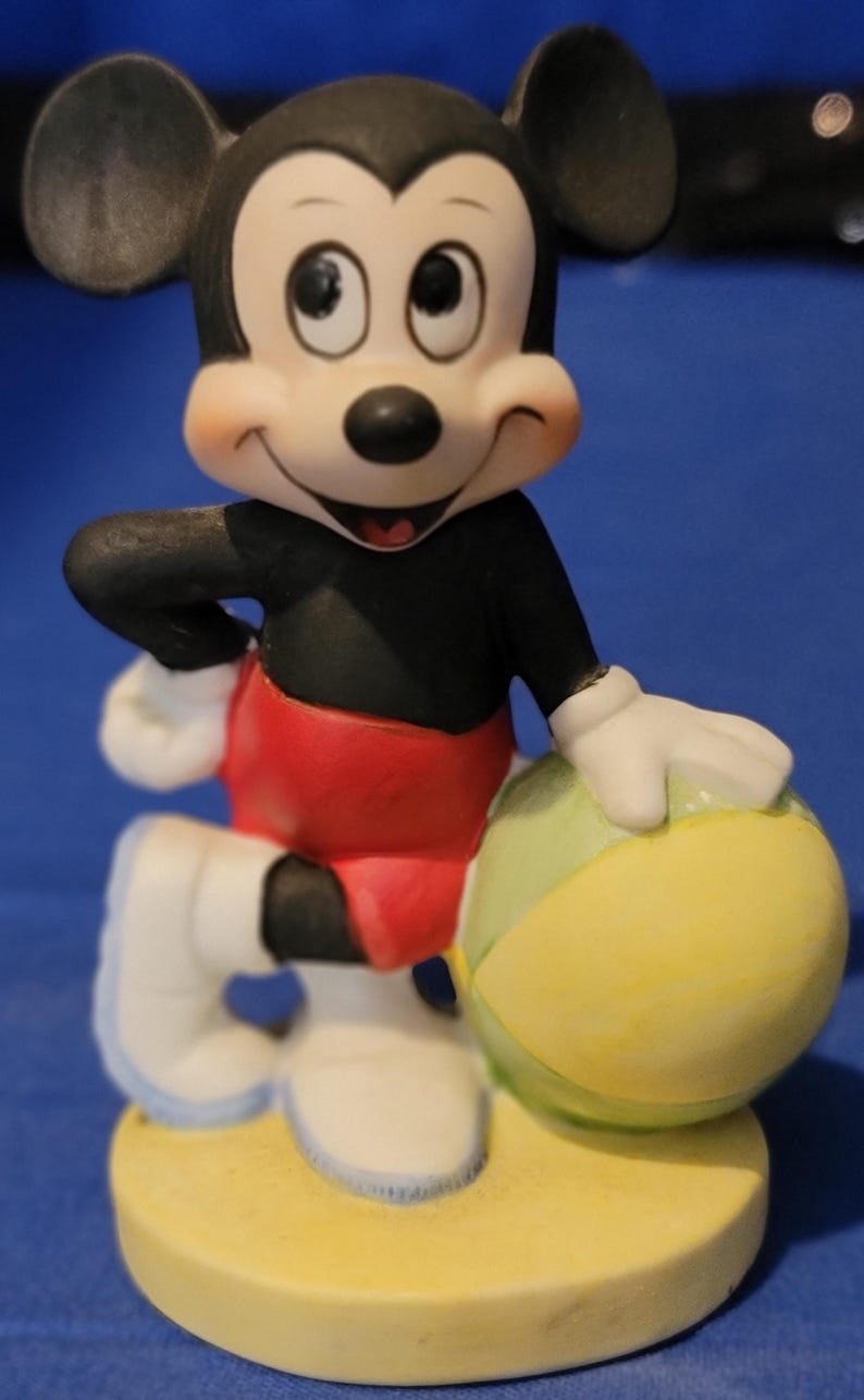 May include: A ceramic figurine of Mickey Mouse, with a black torso, red shorts, and white gloves and shoes. The figure is holding a yellow and green beach ball. The base is yellow.