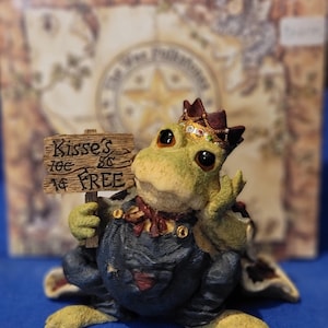 May include: A whimsical frog figurine wearing a crown and holding a sign that reads "Kisses 10¢, 5¢, FREE." The frog is dressed in a dark blue outfit with a red bow tie. The figurine has a detailed, textured appearance.