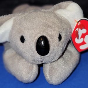 May include: A plush koala bear toy in shades of gray with white accents on the ears. It has black eyes and a black nose. A red heart-shaped tag with a yellow star is attached.