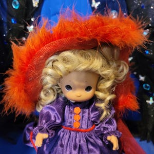 May include: A Precious Moments doll wearing a purple dress with orange buttons and trim. The doll has blonde curly hair and is wearing a red hat with orange feathers. The background is blue and black.