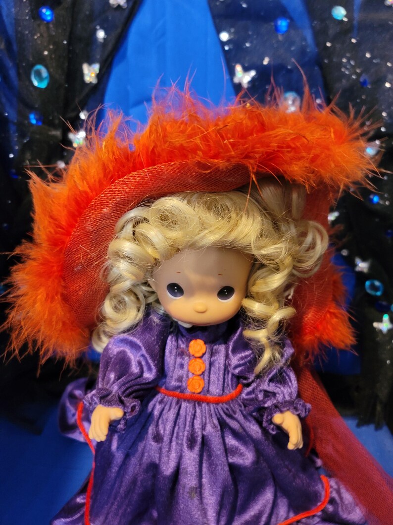 May include: A Precious Moments doll wearing a purple dress with orange buttons and trim. The doll has blonde curly hair and is wearing a red hat with orange feathers. The background is blue and black.