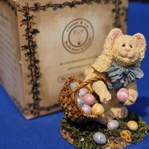 May include: A cream-colored bunny figurine with a blue bow tie, holding a basket of pastel Easter eggs. The figurine is on a grassy base with scattered eggs. A cardboard box with text is in the background.