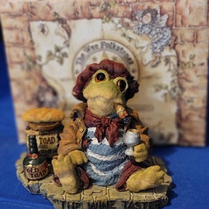 May include: A whimsical figurine of a frog dressed as a wine taster, wearing a beret, scarf, and jacket, holding a wine glass and a cigar. The base reads "THE WINE TASTER". A bottle of "OLD TOAD" wine is also present.