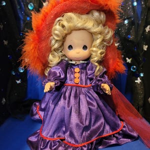 May include: A Precious Moments doll with blonde curly hair, wearing a purple satin dress with orange trim and buttons. The doll has a matching red hat with orange feathers. The doll is a collectible item.