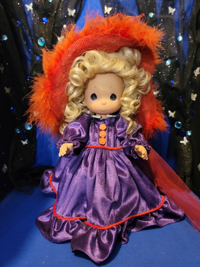 May include: A Precious Moments doll with blonde curly hair, wearing a purple satin dress with orange trim and buttons. The doll has a matching red hat with orange feathers. The doll is a collectible item.