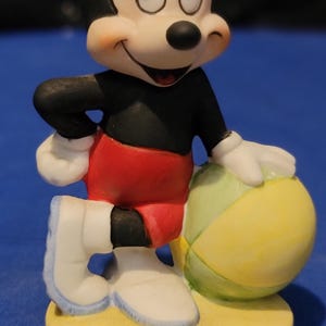 May include: A ceramic Mickey Mouse figurine. The figure wears a black top, red shorts, white gloves and shoes, and is holding a yellow and green beach ball. It stands on a yellow base. The figurine has a smiling expression.