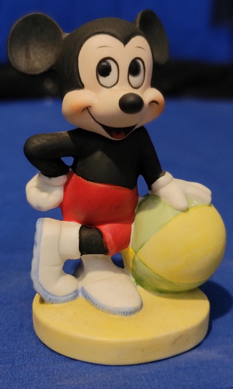 May include: A ceramic Mickey Mouse figurine. The figure wears a black top, red shorts, white gloves and shoes, and is holding a yellow and green beach ball. It stands on a yellow base. The figurine has a smiling expression.