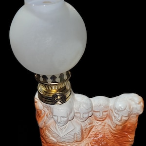 May include: A decorative oil lamp featuring a white frosted glass globe and a brass-colored metal base. The base is sculpted with the faces of four figures, painted in shades of white and orange. The text "MoneySavingDeals58" is visible in the image.