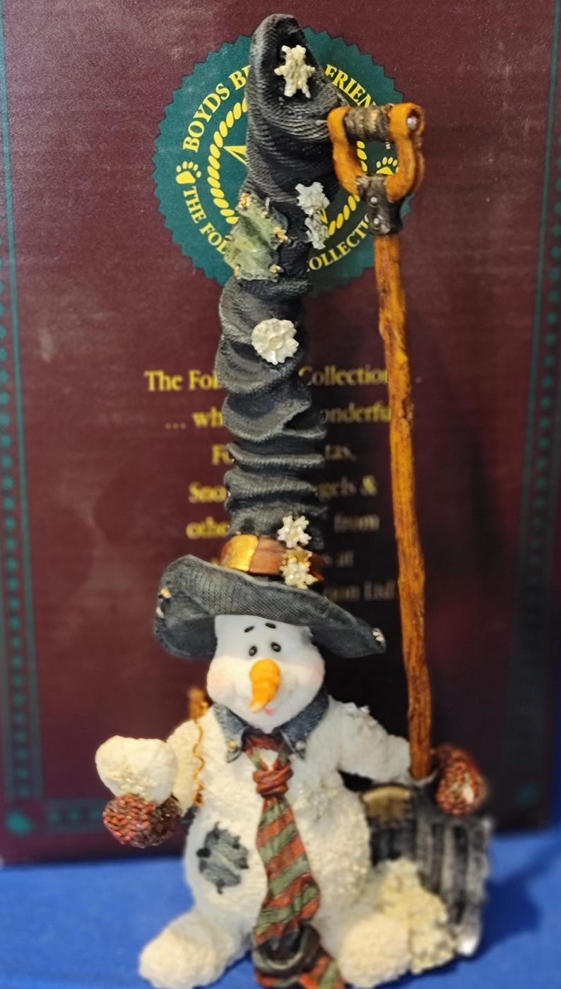 K&ouml;nnte beinhalten: Eine skurrile Schneemannfigur aus der Boyds Collection, die einen schwarzen Hut mit Sternen tr&auml;gt und einen Besen h&auml;lt. Der Schneemann hat eine Karottennase, eine gemusterte Krawatte und einen geflickten Mantel. Die Figur ist ein Sammlerst&uuml;ck.