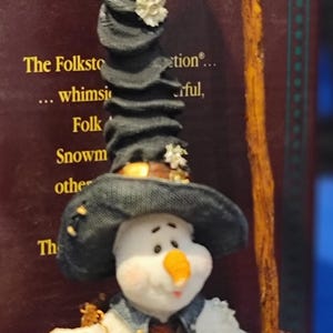 K&ouml;nnte beinhalten: Eine skurrile Schneemannfigur aus der Boyds Bears-Kollektion. Der Schneemann tr&auml;gt einen schwarzen Hut, eine Jeansjacke und einen gestreiften Schal. Er h&auml;lt eine Schaufel und einen kleinen, runden Gegenstand. Die Figur hat einen fr&ouml;hlichen Ausdruck.