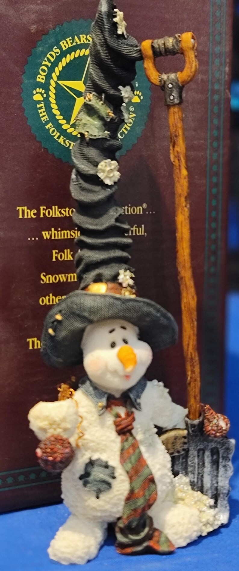 K&ouml;nnte beinhalten: Eine skurrile Schneemannfigur aus der Boyds Bears-Kollektion. Der Schneemann tr&auml;gt einen schwarzen Hut, eine Jeansjacke und einen gestreiften Schal. Er h&auml;lt eine Schaufel und einen kleinen, runden Gegenstand. Die Figur hat einen fr&ouml;hlichen Ausdruck.