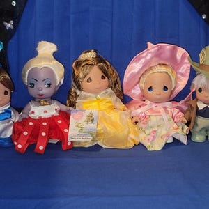 May include: Five Precious Moments figurines of Disney characters, including Belle, the Evil Queen, and the Mad Hatter. The dolls are dressed in colorful outfits, with Belle in a blue and white dress and the Evil Queen in a red and white polka dot dress.
