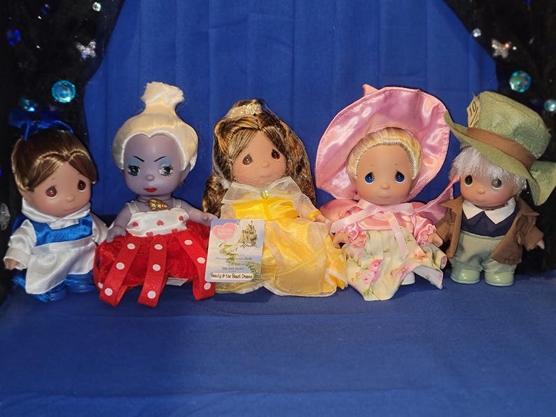 May include: Five Precious Moments figurines of Disney characters, including Belle, the Evil Queen, and the Mad Hatter. The dolls are dressed in colorful outfits, with Belle in a blue and white dress and the Evil Queen in a red and white polka dot dress.