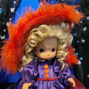 May include: A Precious Moments doll with blonde curly hair, wearing a purple dress with orange flower-shaped buttons and a red belt. The doll has a red hat with orange feathers.