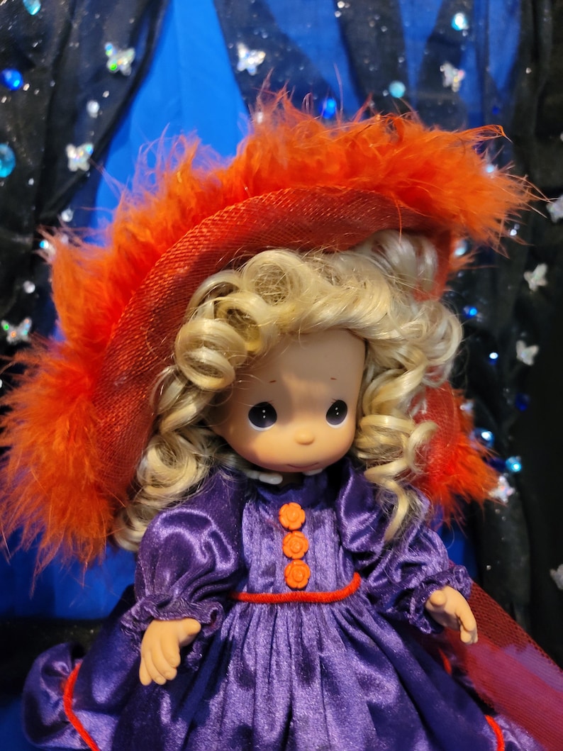 May include: A Precious Moments doll with blonde curly hair, wearing a purple dress with orange flower-shaped buttons and a red belt. The doll has a red hat with orange feathers.