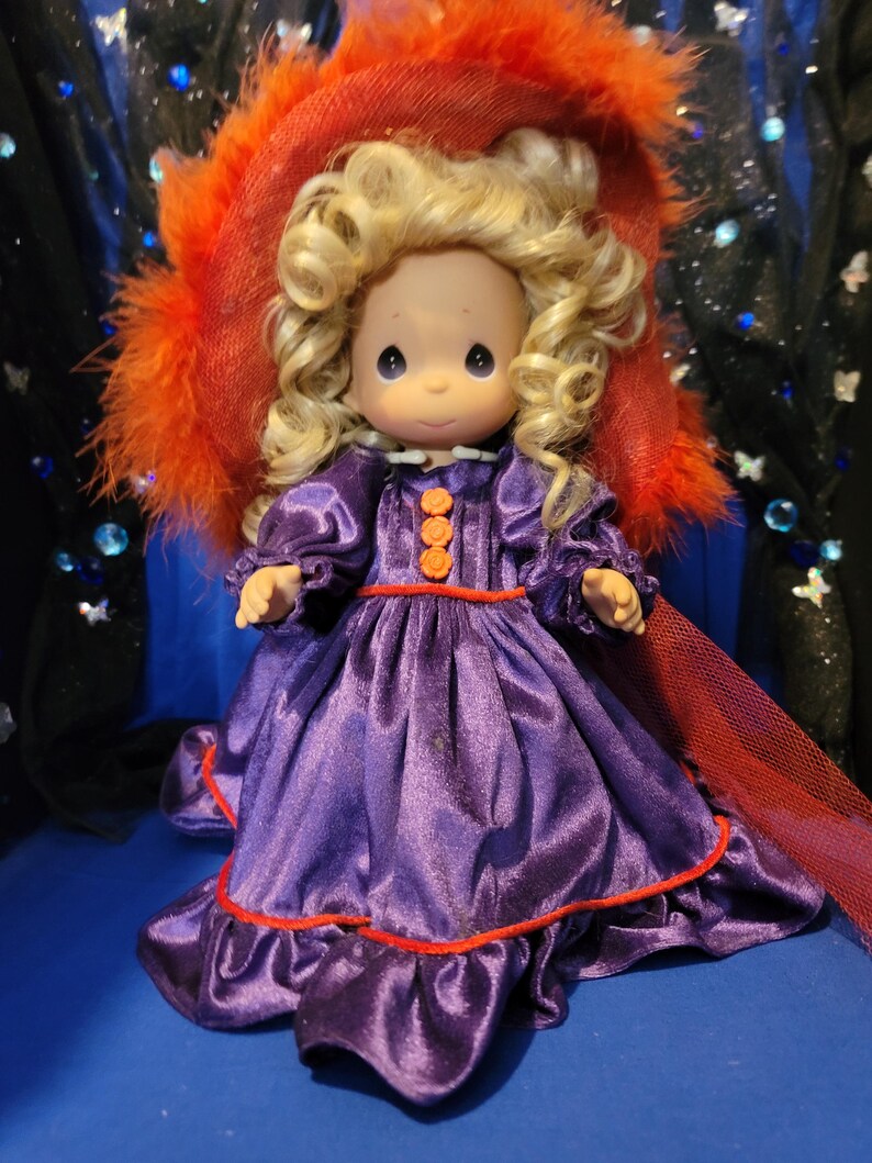 May include: A Precious Moments figurine of a girl with blonde curly hair, wearing a purple satin dress with orange buttons and trim. She is wearing a red hat with orange feathers. The doll has a sweet expression.