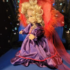 May include: A vintage doll with blonde curly hair, wearing a purple satin dress with red trim and a matching hat adorned with orange feathers. The doll is posed against a dark blue backdrop.