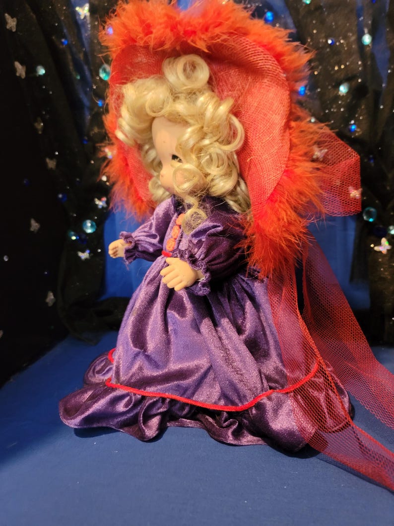 May include: A vintage doll with blonde curly hair, wearing a purple satin dress with red trim and a matching hat adorned with orange feathers. The doll is posed against a dark blue backdrop.