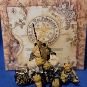 May include: A figurine of a frog dressed in a white shirt and dark blue pants, holding a fishing rod. A smaller frog figurine sits beside it. The background features a box with the text "The Wee Folkstones & Friends".