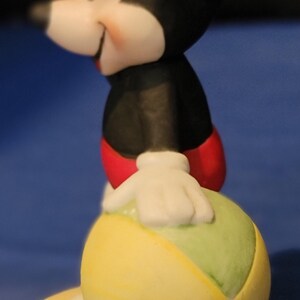 May include: A ceramic Mickey Mouse figurine, dressed in a black jacket, red shorts, and white gloves. He is holding a yellow, green, and white ball. The figurine stands on a yellow base. The background is a solid blue.
