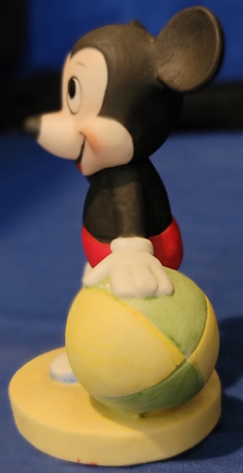 May include: A ceramic Mickey Mouse figurine, dressed in a black jacket, red shorts, and white gloves. He is holding a yellow, green, and white ball. The figurine stands on a yellow base. The background is a solid blue.
