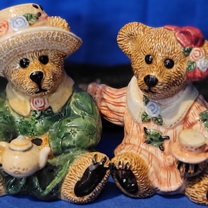 May include: Two ceramic teddy bear figurines. One bear wears a green jacket and hat, holding a teapot. The other bear wears a pink and white striped dress and a hat. Both bears have black eyes and paws.