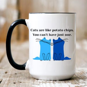 May include: White ceramic mug with a black handle and rim. The mug features a blue and dark blue cat illustration and the text: "Cats are like potato chips. You can't have just one."