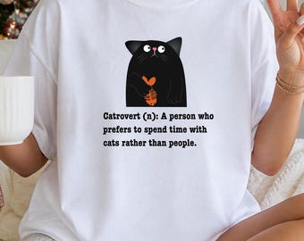 May include: A white t-shirt featuring a graphic of a black cat holding an orange fish skeleton. Below the cat, the text reads: Catrovert (n): A person who prefers to spend time with cats rather than people. The shirt has a crew neckline and short sleeves.