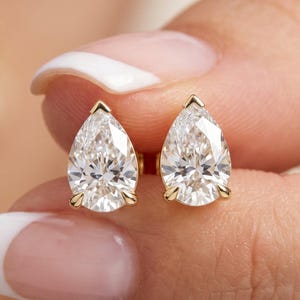 Può includere: Un paio di orecchini a lobo con diamanti a forma di pera con montatura in oro. I diamanti sono trasparenti e sfaccettati, riflettendo la luce. Gli orecchini sono tenuti in mano da una persona con la pelle chiara.