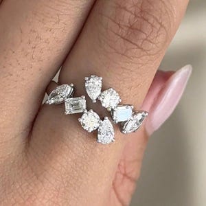 Può includere: Un anello d'argento con un design floreale, caratterizzato da molteplici pietre preziose chiare e scintillanti di varie forme. L'anello è indossato su un dito con smalto rosa chiaro.