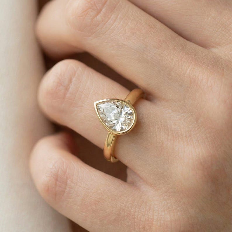 May include: A gold ring featuring a pear-shaped diamond. The diamond is set in a bezel setting, and the band is a smooth, polished gold. The ring is shown on a person's finger, with the hand in the background.